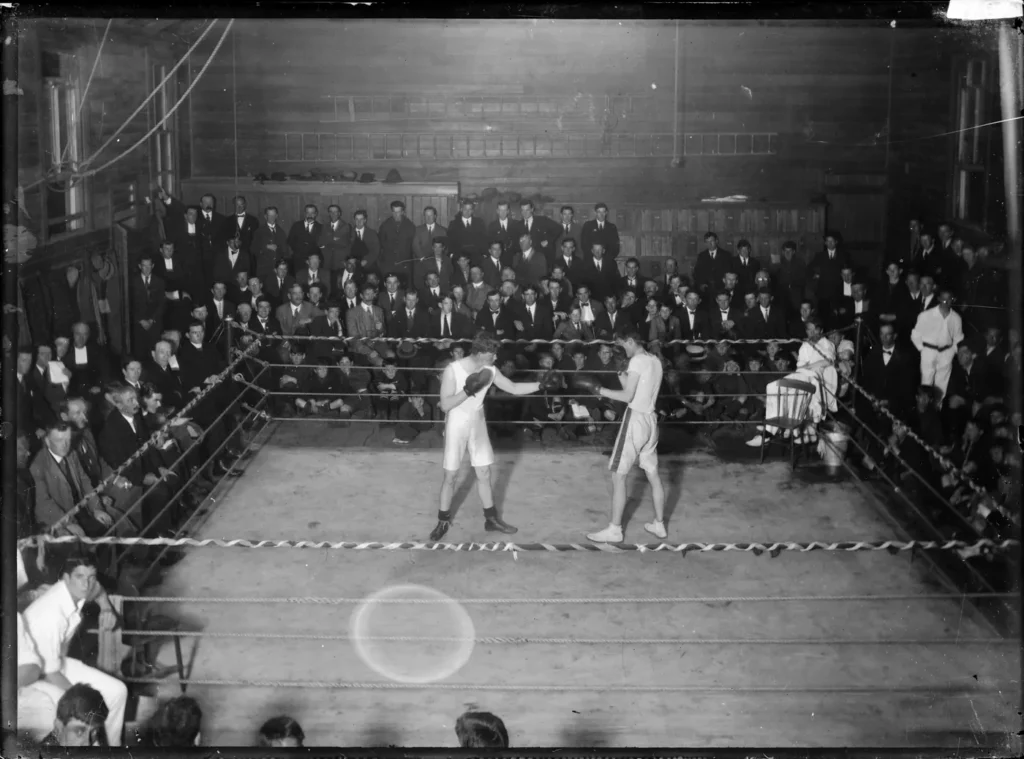 Photographie ancienne en noir et blanc : deux boxeurs dans un ring entouré d’une foule d’hommes en costume