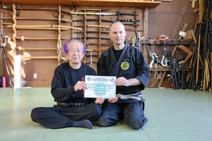 Masaaki Hatsumi, assis en tenue sombre, remet un diplôme à un élève dans un dojo dont les murs portent des armes de bois