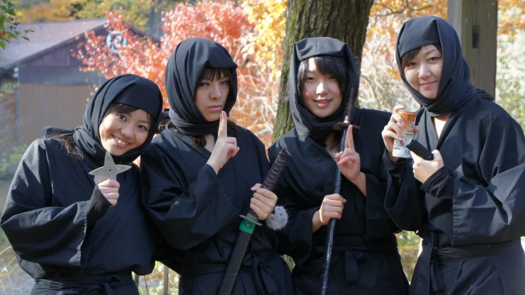Quatre visiteuses en costume noir de ninja posent en riant devant les érables d’automne, à Kōka
